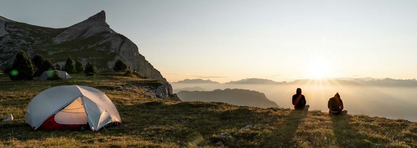 Zwei Personen sitzen vor ihrem Zelt und betrachten den Sonnenuntergang vor einer beleuchteten Bergkulisse.