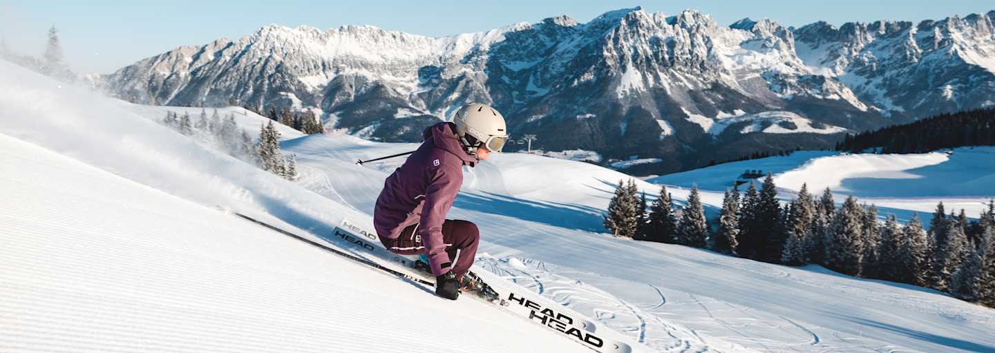 Eine Skifahrerin carvt im Skigebiet Wilder Kaiser- Brixental über eine frisch präparierte Piste. Im Hintergrund die markante Bergkulisse des Wilden Kaisers.