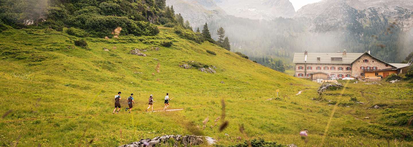 Vier Trailrunner laufen entlang einer Wiese auf einem Berg in Richtung einer Hütte mit roten Fensterläden.