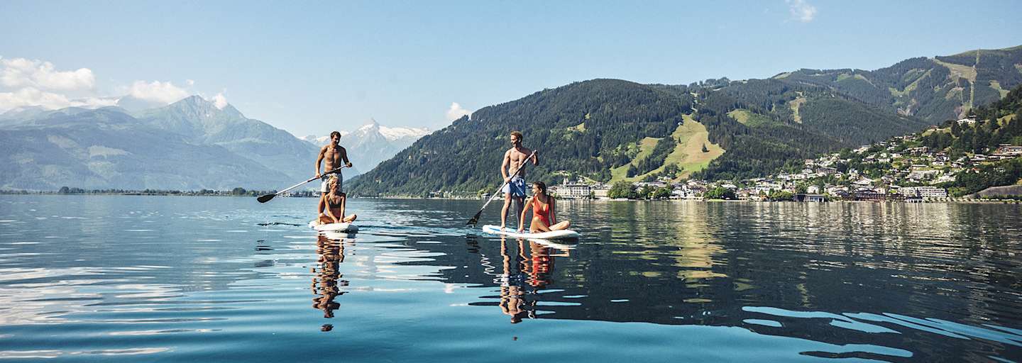 Zwei Männer tauche mit einem Puddle ihr Stand-up-paddeln an. Jeweils eine Frau, sitz am vorderen Ende. Sie gleiten über denn Zeller See. Hinter ihnen ein Bergpanorama.