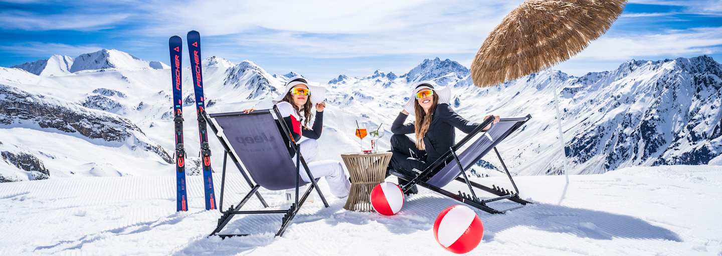 Zwei Frauen sitzen auf Sonnenliegestühlen in einer Winterlandschaft und lächeln in die Kamera.