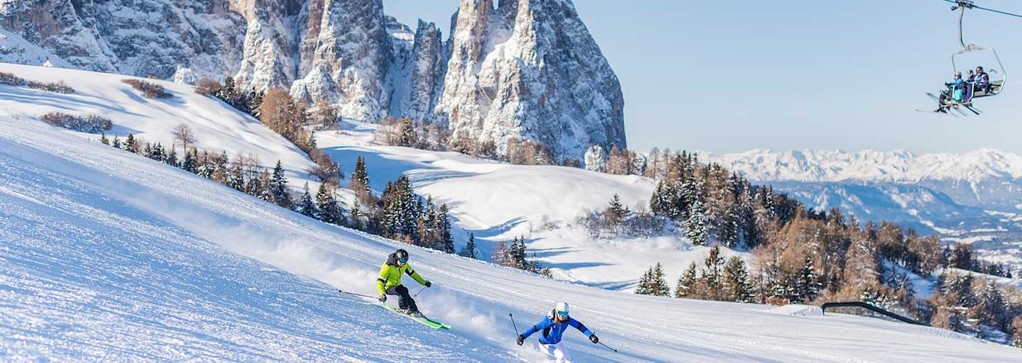 Zwei Skifahrer fahren eine frisch präparierte Piste in der Dolomitenregion Seiser Alm hinunter. Im Hintergrund ist das verschneite Bergpanorama zu sehen.