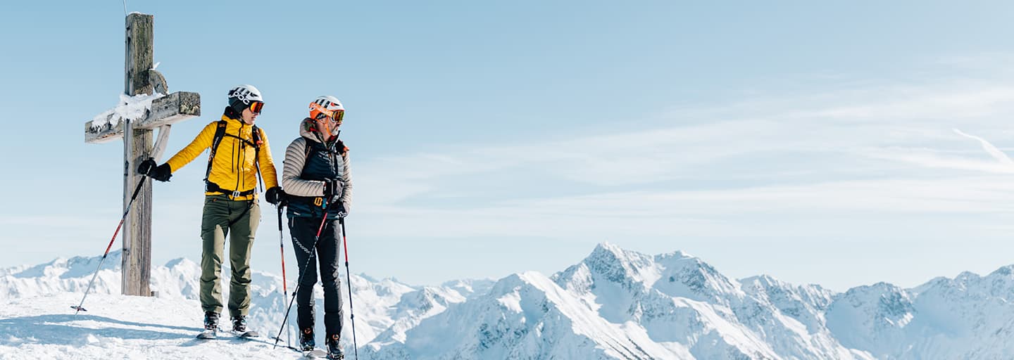 Eine Frau und ein Mann stehen neben dem Gipfelkreuz auf Tourenskiern und blicken über die verschneite Berglandschaft.