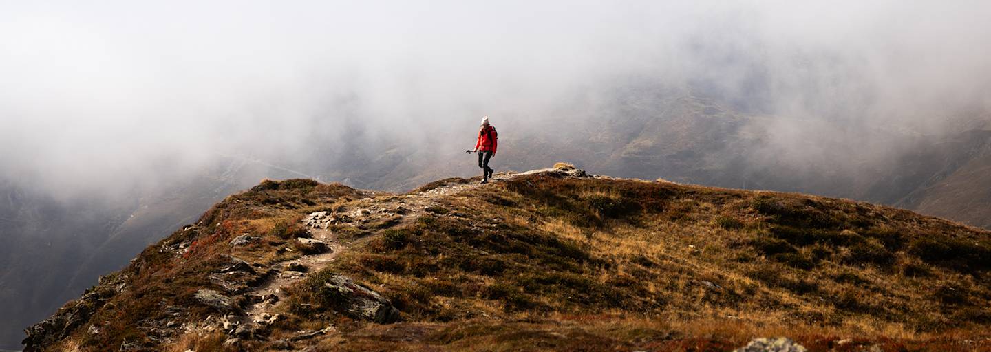 Person wandert in roter Outdoor-Jacke auf einem Bergpfad durch eine neblige Gebirgslandschaft.