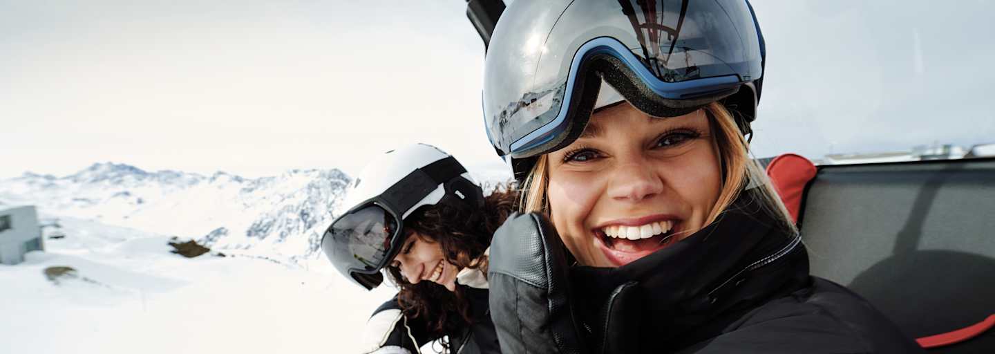 Eine Frau mit Skibrille und Helm auf dem Kopf sitz in einem Sessellift und lacht uns entgegen. Neben ihr sitzt eine weitere lächelnde Frau.