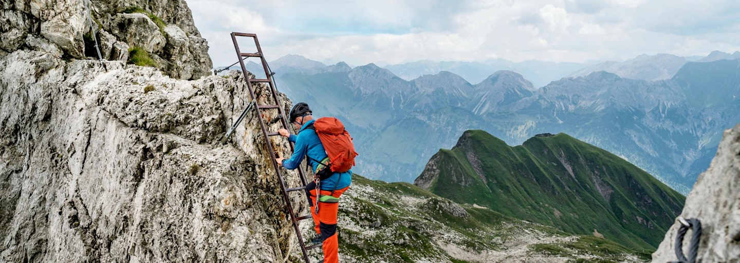 Ein Kletterer nimmt die Stahlleiter, im Hintergrund ist ein Bergsee zu sehen