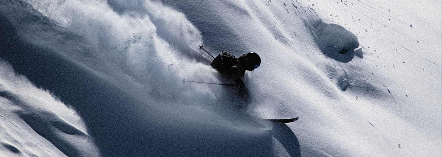 Skifahrer gleitet dynamisch durch unberührten Tiefschnee, wirbelt Pulverschnee auf und meistert eine steile Abfahrt in alpiner Winterlandschaft.