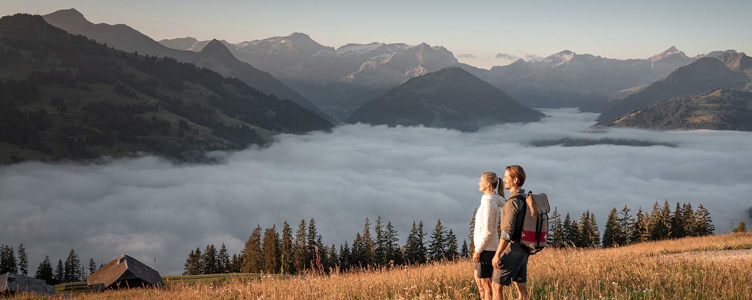 Zwei Wanderer genießen den Blick über ein Nebelmeer im herbstlichen Gstaad mit Bergpanorama im Hintergrund.