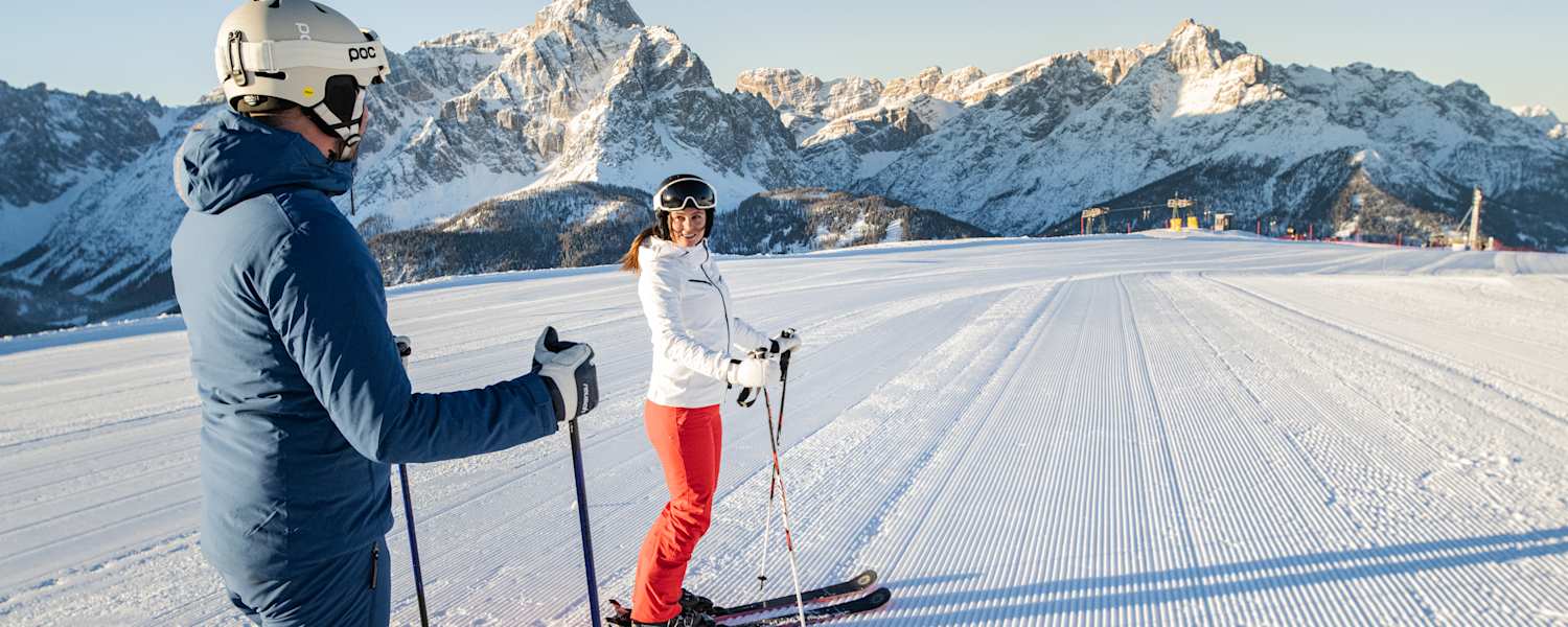 zwei skifahrer stehen auf der piste und schauen sich an, der himmel ist strahlend blau und die schneebedeckten bergspitzen stehen im hintergrund