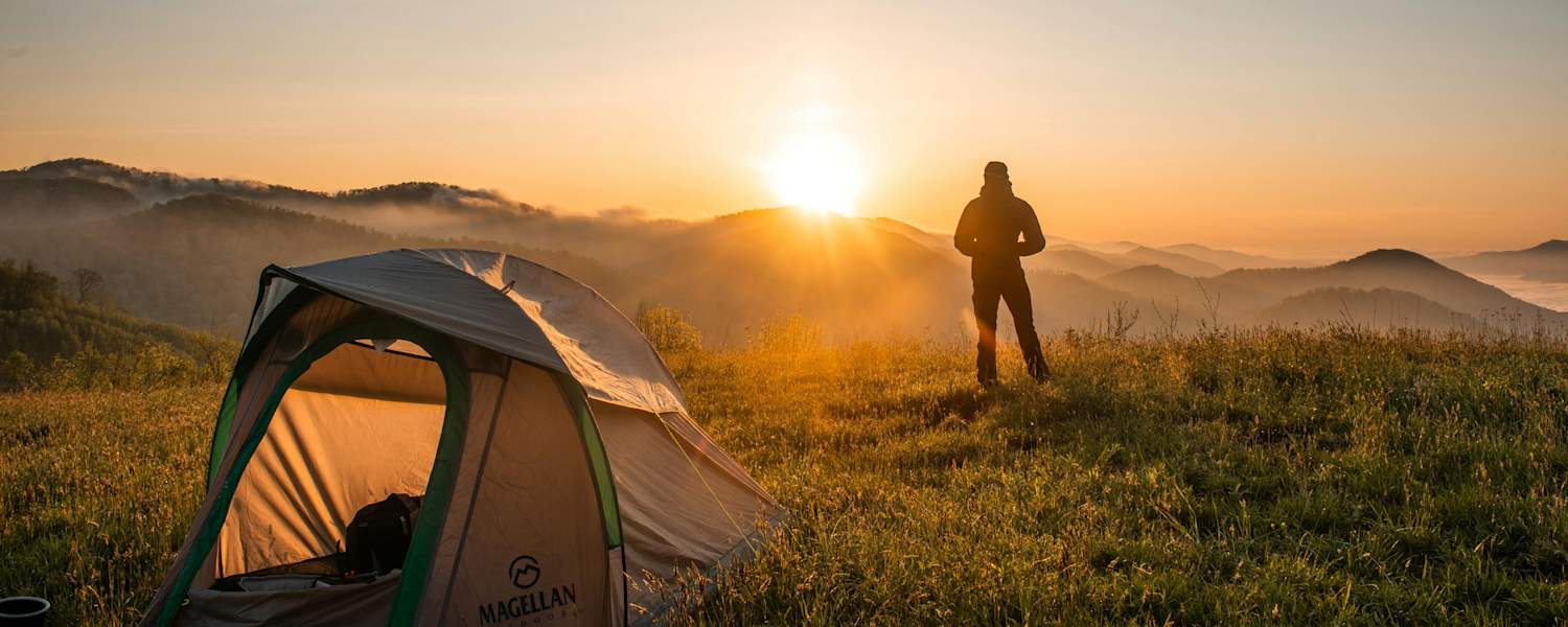 Ein Mann steht vor seinem Zelt und schaut zu wie die Sonne hinter den Bergen untergeht.