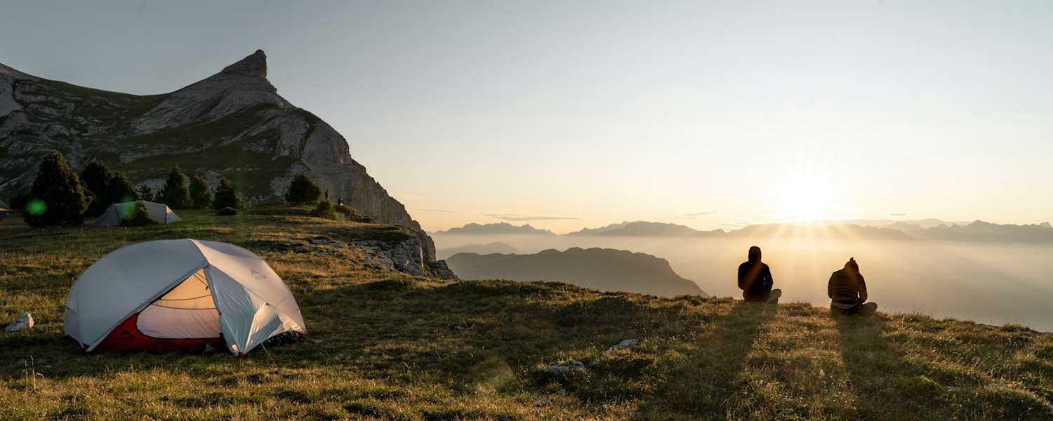 Zwei Personen sitzen vor ihrem Zelt und betrachten den Sonnenuntergang vor einer beleuchteten Bergkulisse.
