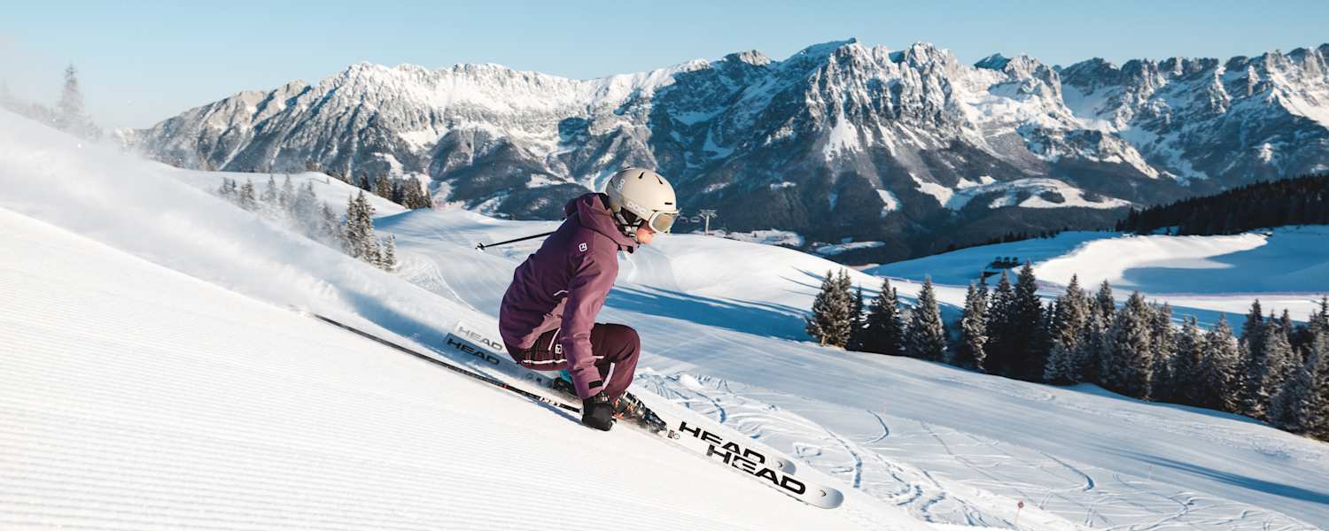 Eine Skifahrerin carvt im Skigebiet Wilder Kaiser- Brixental über eine frisch präparierte Piste. Im Hintergrund die markante Bergkulisse des Wilden Kaisers.