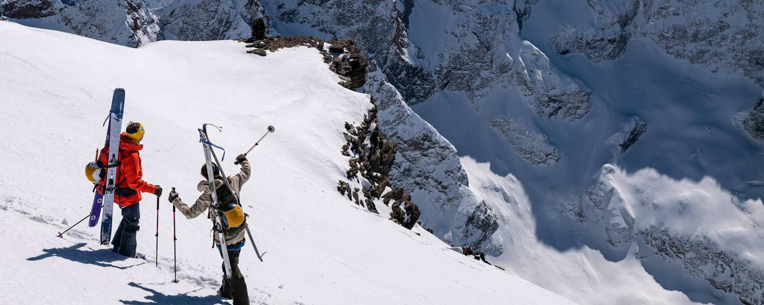 Auf einem schneebedeckten Hang stehen zwei Skitourengeher und zeigen in die Ferne. Sie sind mit der Bergtagen-Kollektion von Fjällräven ausgerüstet.