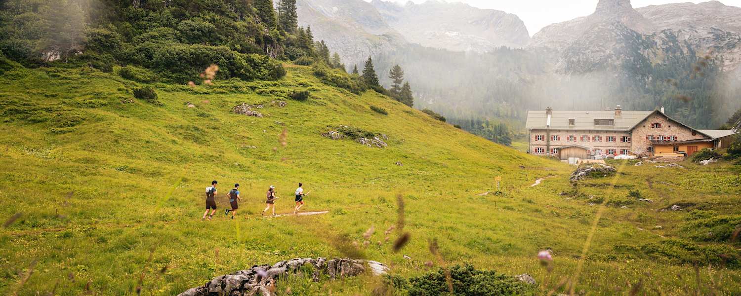 Vier Trailrunner laufen entlang einer Wiese auf einem Berg in Richtung einer Hütte mit roten Fensterläden.
