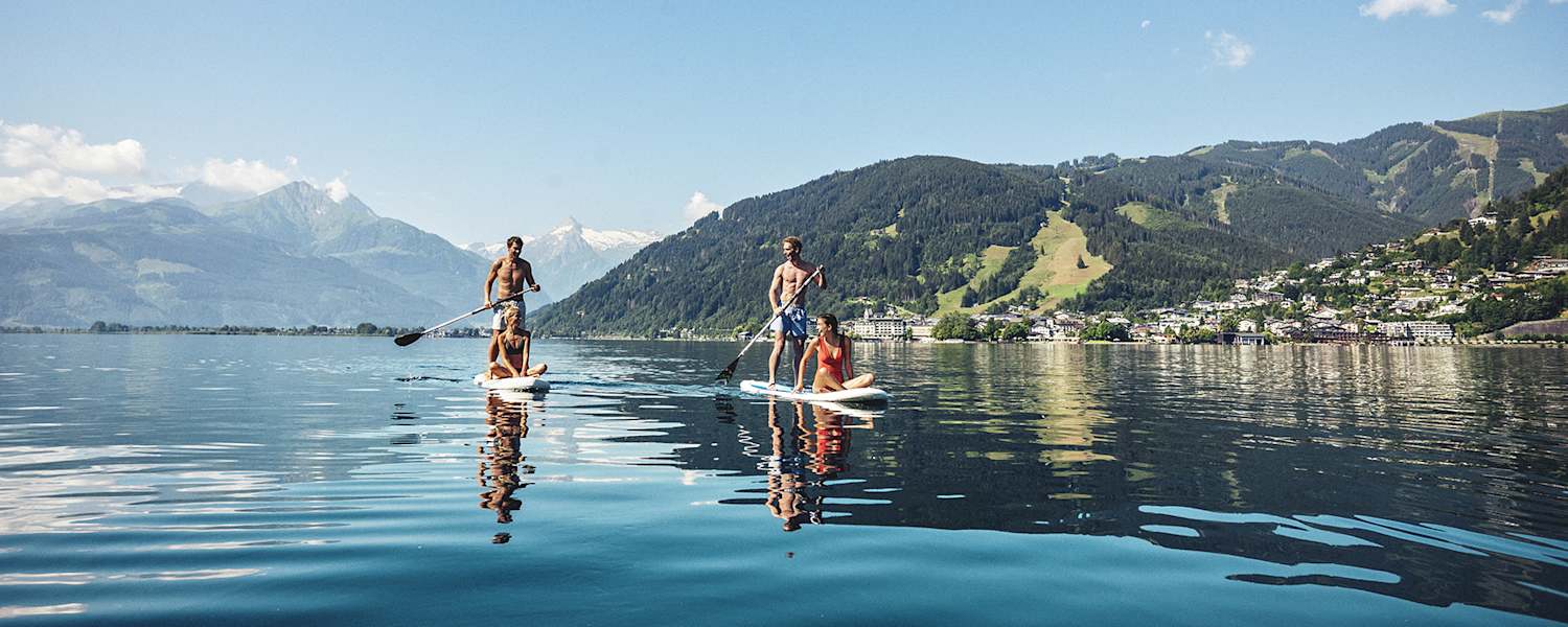 Zwei Männer tauche mit einem Puddle ihr Stand-up-paddeln an. Jeweils eine Frau, sitz am vorderen Ende. Sie gleiten über denn Zeller See. Hinter ihnen ein Bergpanorama.