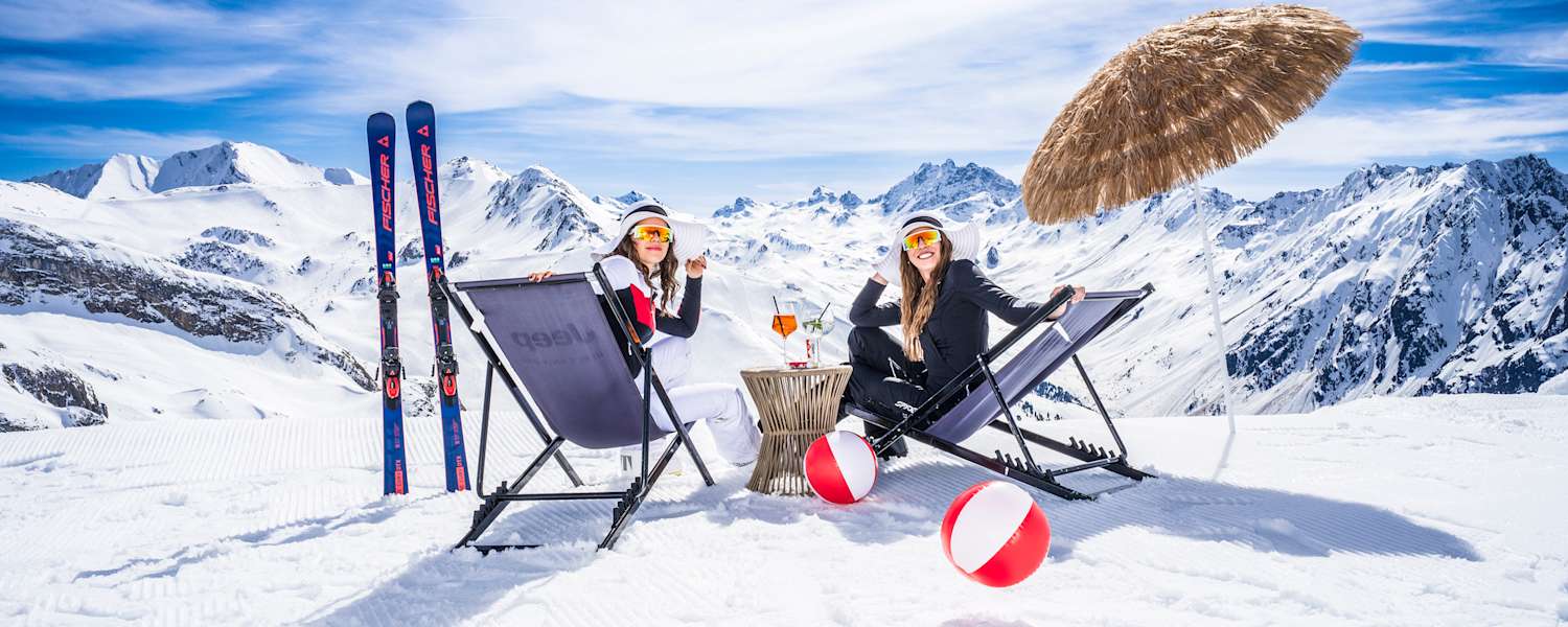 Zwei Frauen sitzen auf Sonnenliegestühlen in einer Winterlandschaft und lächeln in die Kamera.