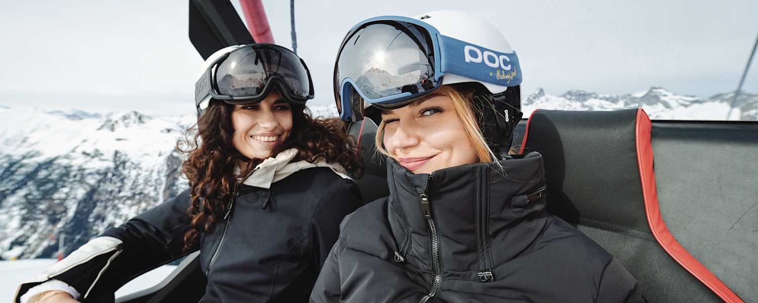 Eine blonde Frau zwinkert in die Kamera. Eine andere Frau, die neben ihr sitzt lächelt ihr dabei zu.