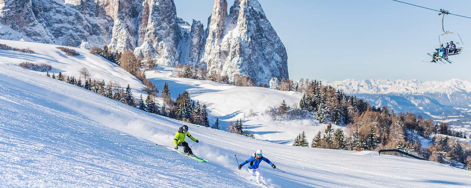 Zwei Skifahrer fahren eine frisch präparierte Piste in der Dolomitenregion Seiser Alm hinunter. Im Hintergrund ist das verschneite Bergpanorama zu sehen.