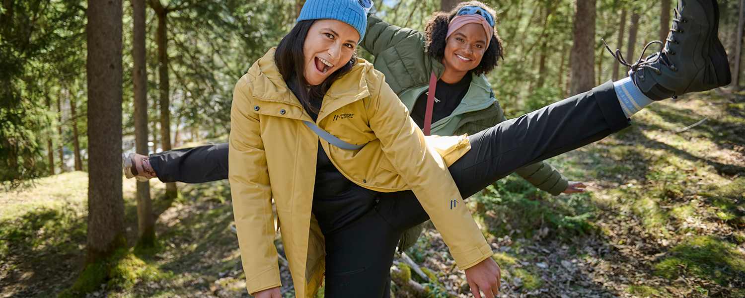 Zwei Personen in moderner Outdoorbekleidung von Maier Sports, aktiv im Wald unterwegs, umgeben von Bäumen und Sonnenlicht.