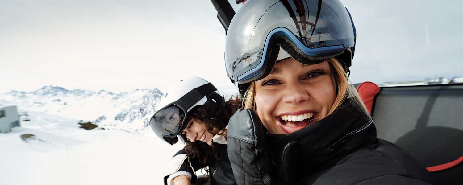 Eine Frau mit Skibrille und Helm auf dem Kopf sitz in einem Sessellift und lacht uns entgegen. Neben ihr sitzt eine weitere lächelnde Frau.
