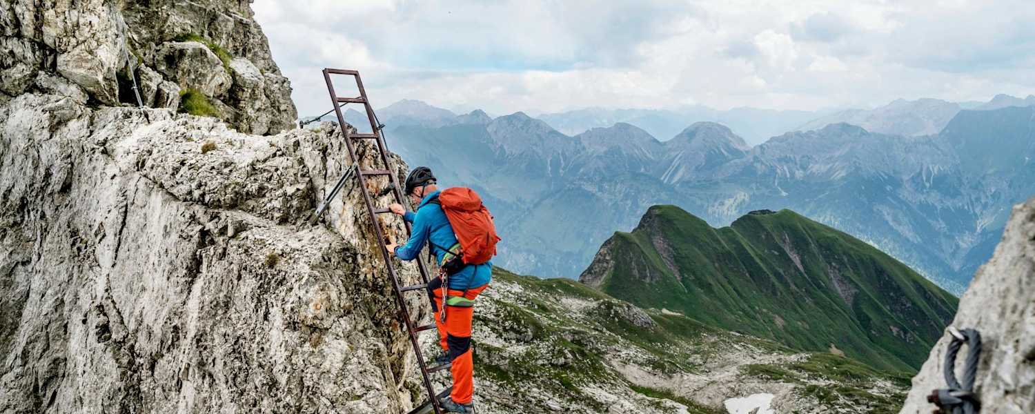 Ein Kletterer nimmt die Stahlleiter, im Hintergrund ist ein Bergsee zu sehen