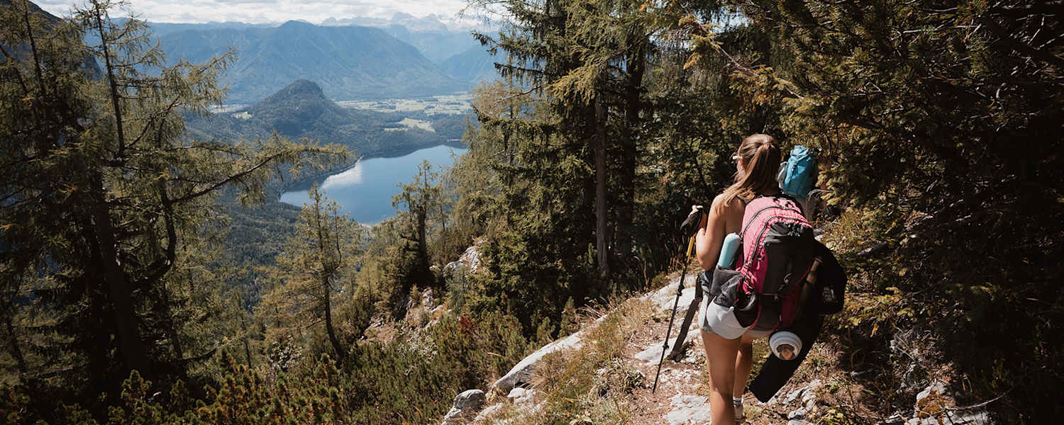 Eine Wandrerin mit Rucksack und Ausrüstung, die durch den Wald auf den Altauseer See hinunterblickt.
