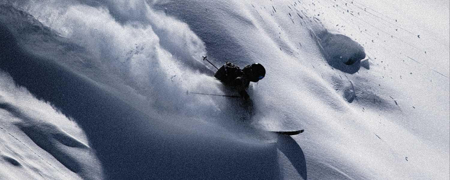 Skifahrer gleitet dynamisch durch unberührten Tiefschnee, wirbelt Pulverschnee auf und meistert eine steile Abfahrt in alpiner Winterlandschaft.