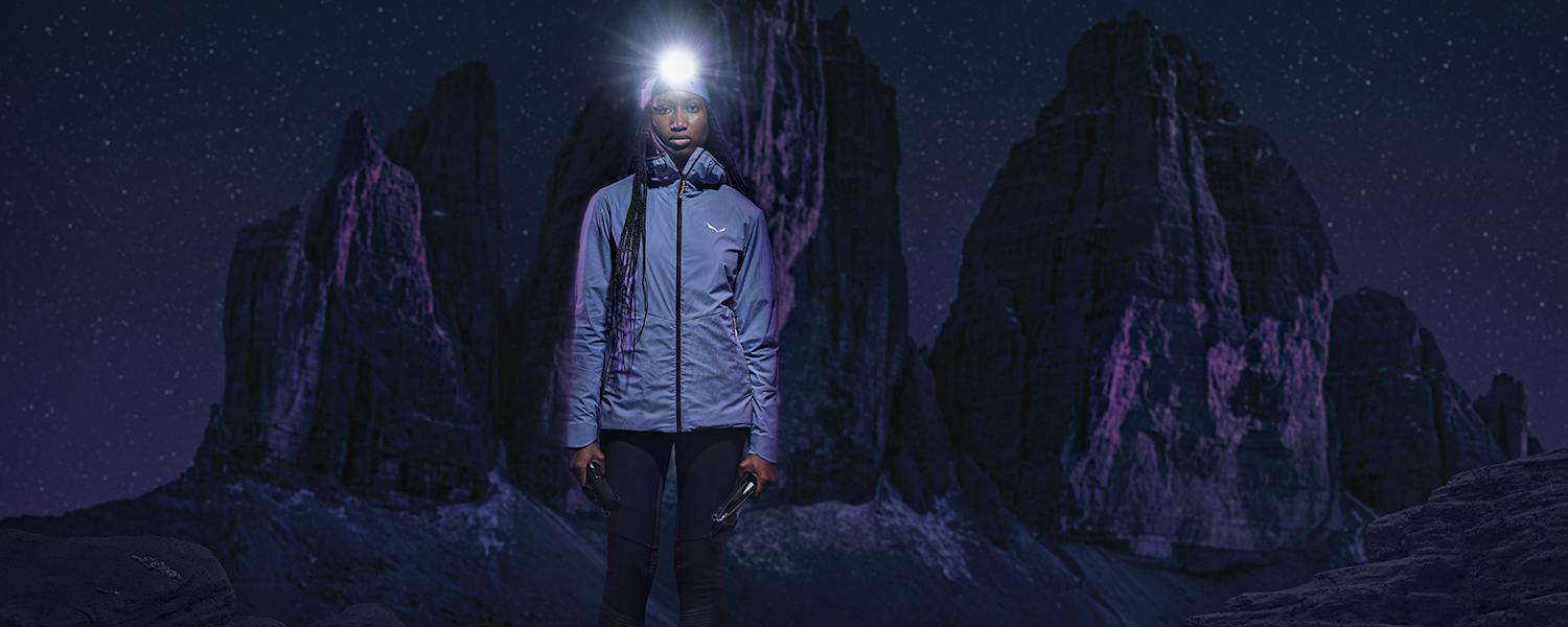 Frau mit Stirnlampe in Speedhiking Outfit von Salewa in der Nacht, im Hintergrund Berge in der Dunkelheit.