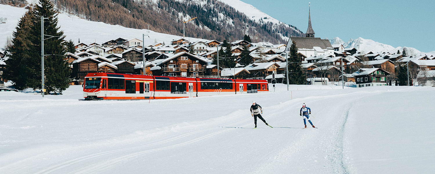 Ein Tal mit 2 Langläufern, einer Bahn und verschneite Tannen.