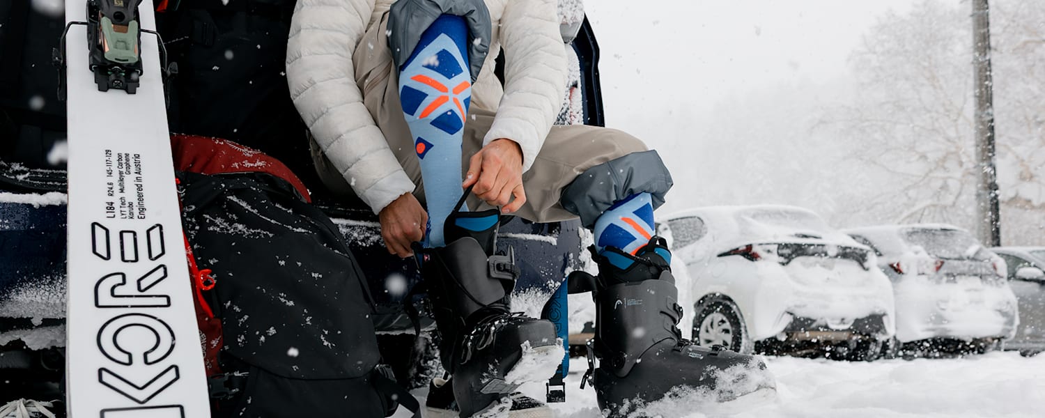 Ein Skifahrer sitzt am Parkplatz und zieht sich im Schnee seine Skischuhe an.