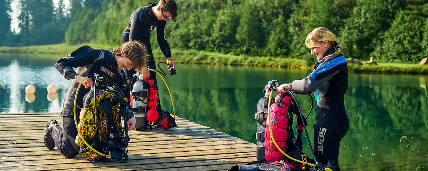 Drei Taucher in Neoprenanzügen knien auf einem Holzsteg am Grüblsee