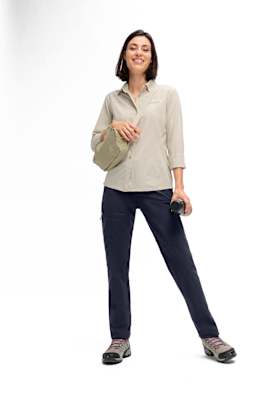 Frau in Maier Sports Outdoorbekleidung mit nachhaltiger Wanderhose und beigem Funktionshemd, kombiniert mit Wanderschuhen und Umhängetasche, vor weißem Hintergrund.