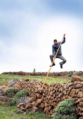 Ein junger Mann springt mit Hilfe eines Holzstabes über eine alte Mauer