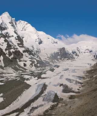 Was für ein Auftakt! Der Sprung ins Abenteuer Alpe-Adria-Trail könnte spektakulärer nicht sein: Schon die ersten Meter führen uns hinab zu den Ausläufern der Pasterze. Blickt man vom Ausgangspunkt ins Tal hinab, kann man die Ausmaße erahnen, die diese Eiszunge einmal hatte. Die Erderwärmung ließ den Gletscher in den letzten 150 Jahren beinahe auf die Hälfte seiner einstigen Größe zusammenschmelzen.