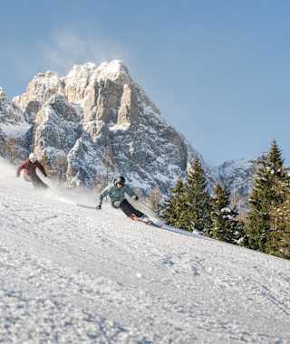 Zwei Skifahren carven in hohem Tempo über eine Skipiste. Im Hintergrund ein schroffer Dolomitengipfel