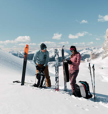 Eine Frau und ein Mann stehen im Schnee auf einem Berg und machen sich für das Skifahren bereit.