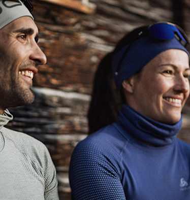 Zwei Personen in funktionaler Winterbekleidung von Odlo, genießen eine Pause im Freien vor einer rustikalen Holzhütte.