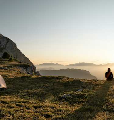 Zwei Personen sitzen vor ihrem Zelt und betrachten den Sonnenuntergang vor einer beleuchteten Bergkulisse.