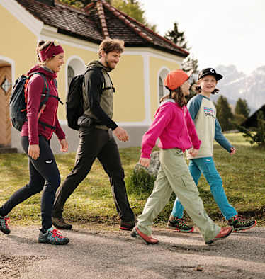 Eine Frau wandert mit einem Mann und zwei Kindern lächelnd durch eine bergige Landschaft im Frühling.