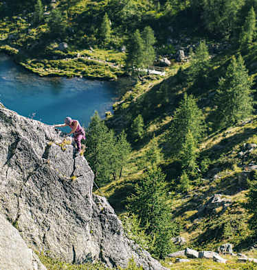 Eine Frau klettert auf einer Felswand entlang, im Hintergrund ist eine sommerliche, grüne Landschaft mit einem See zu sehen.