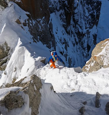 Ein Bergsteiger mit blauer Jacke und orangefarbener Hose erklimmt einen Grat. Dabei ist er mit einem Seil gesichert.