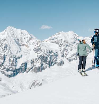 Ein Skifahrer und eine Skifahrerin stehen auf einem Hang und genießen das verschneite Bergpanorama im Ortlergebiet.