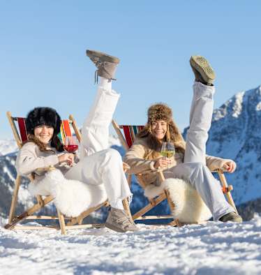 Zwei Frauen sitzen auf Liegestühlen auf einem Berg oben und heben jeweils einen Fuß in die Höhe.