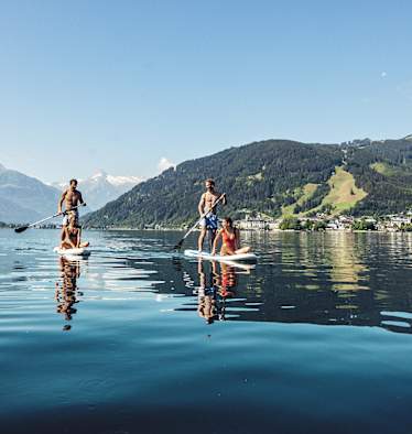 Zwei Männer tauche mit einem Puddle ihr Stand-up-paddeln an. Jeweils eine Frau, sitz am vorderen Ende. Sie gleiten über denn Zeller See. Hinter ihnen ein Bergpanorama.
