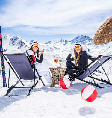 Zwei Frauen sitzen auf Sonnenliegestühlen in einer Winterlandschaft und lächeln in die Kamera.