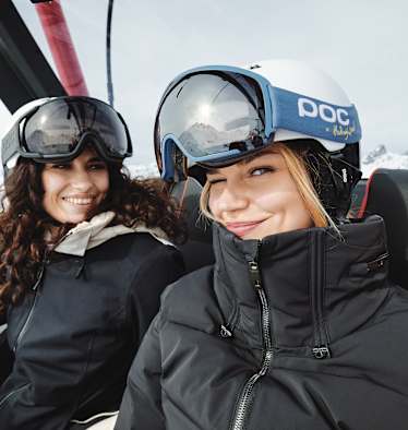 Eine blonde Frau zwinkert in die Kamera. Eine andere Frau, die neben ihr sitzt lächelt ihr dabei zu.