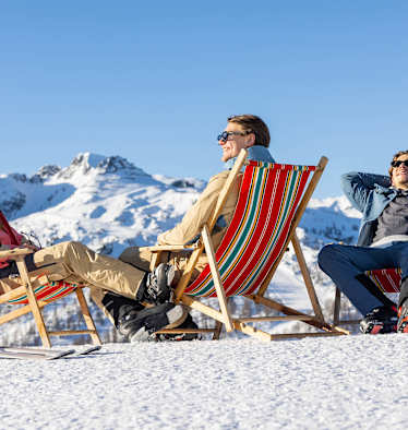 Eine Frau und zwei Männer sitzen in roten Liegenstühlen auf einem verschneiten Berg und genießen die Sonnestrahlen.