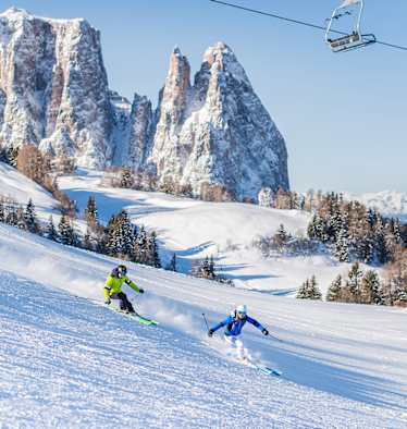 Zwei Skifahrer fahren eine frisch präparierte Piste in der Dolomitenregion Seiser Alm hinunter. Im Hintergrund ist das verschneite Bergpanorama zu sehen.