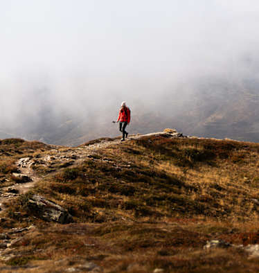 Person wandert in roter Outdoor-Jacke auf einem Bergpfad durch eine neblige Gebirgslandschaft.