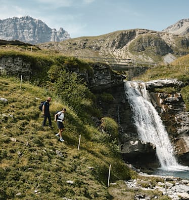 Zwei Frauen wandern einen Hügel hinab. Neben ihnen verläuft ein kleiner Wasserfall und im Hintergrund ist eine alpine Landschaft sichtbar.